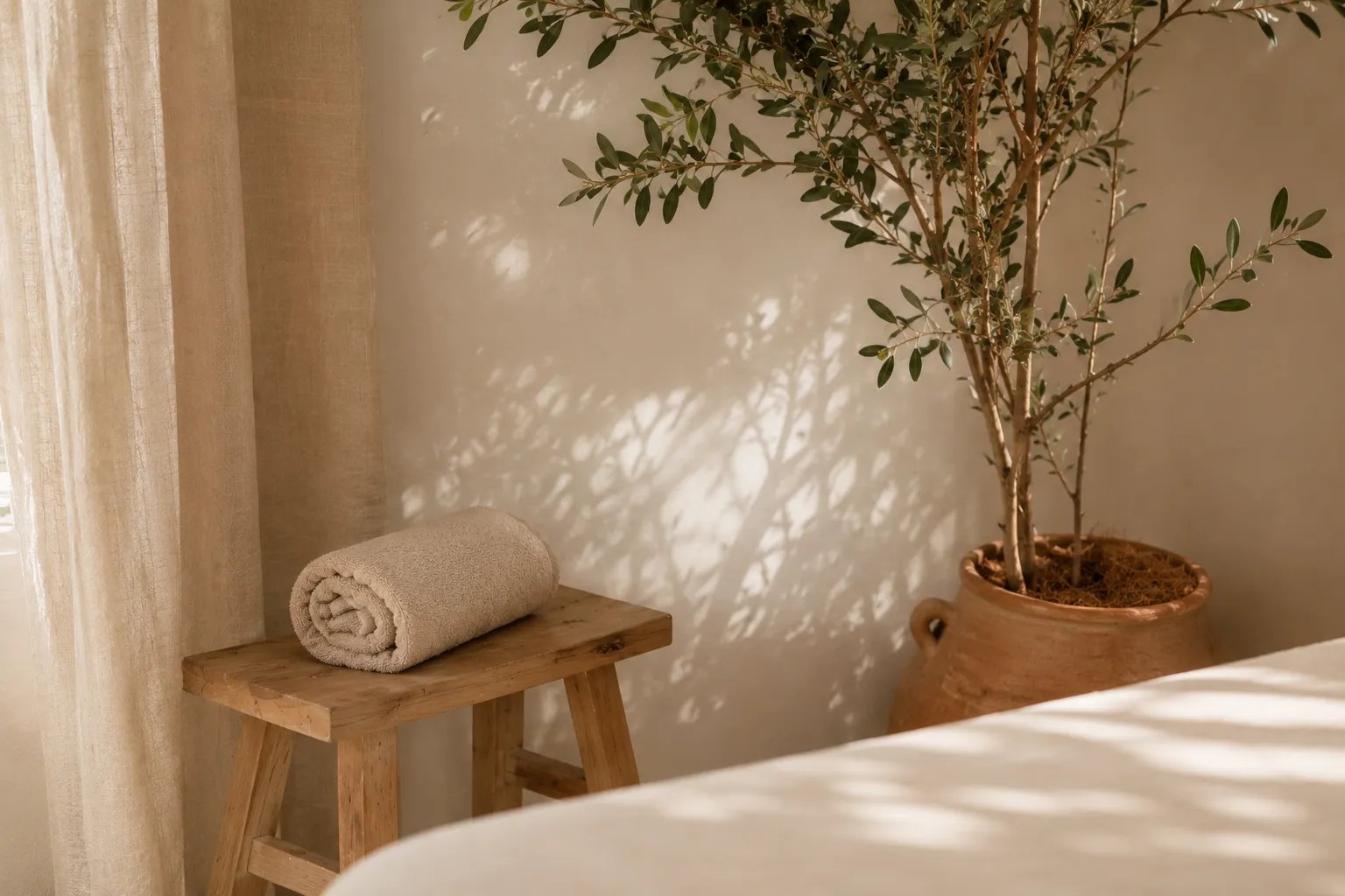 Coin d'une salle de soin : serviette roulée sur un tabouret en bois, olivier en pot et ombres de feuillage sur un mur crème
